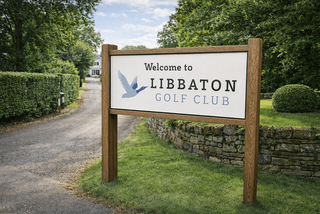 A driveway with stone wall, grass verges and trees featuring a large a twin posted Welcome to Libbaton Golf Club entrance sign